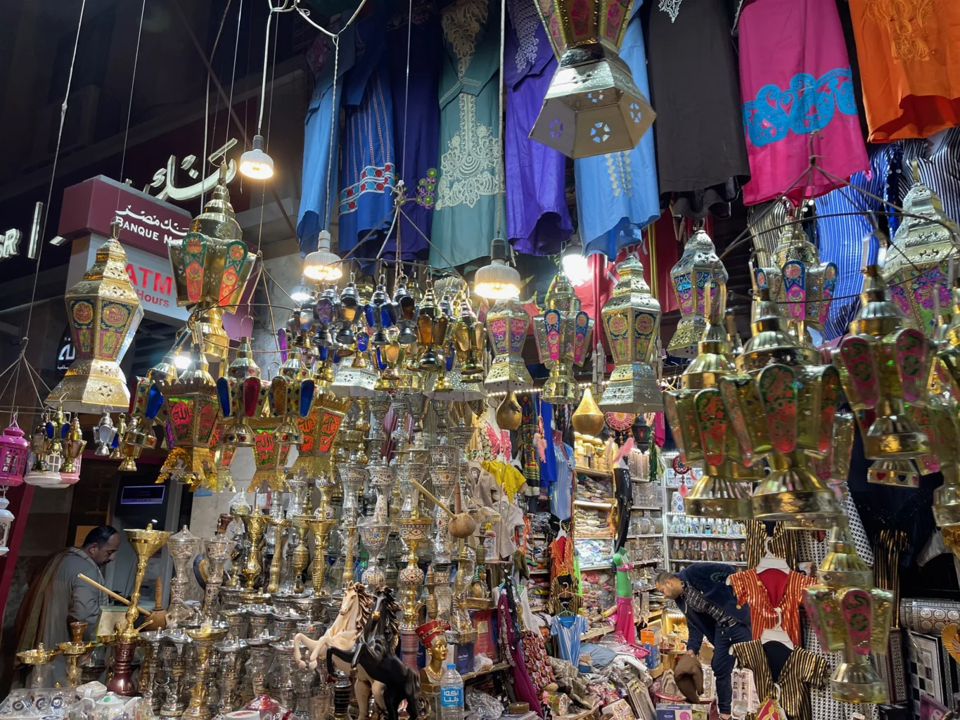 The historic Khan el-Khalili bazaar in Cairo with an abundant variety of market choices. Two men are seen in the opulent market of a densely packed market stall with traditional Egyptian handicrafts. Photo Credits: Celena Brown 2022.
